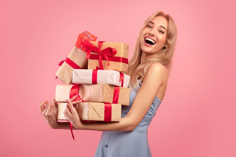 A blonde woman in her 20s holds a pile of wrapped gifts.