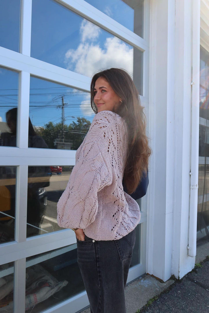 Woman wearing a beige knitted sweater standing in front of a glass door with reflections.